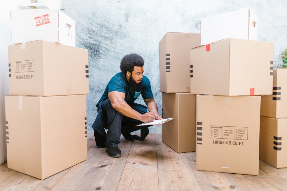 A man standing indoors among numerous cardboard moving boxes in various sizes, some with red and black packing tape and labels, on a concrete or textured wall background. He is casually dressed in an 