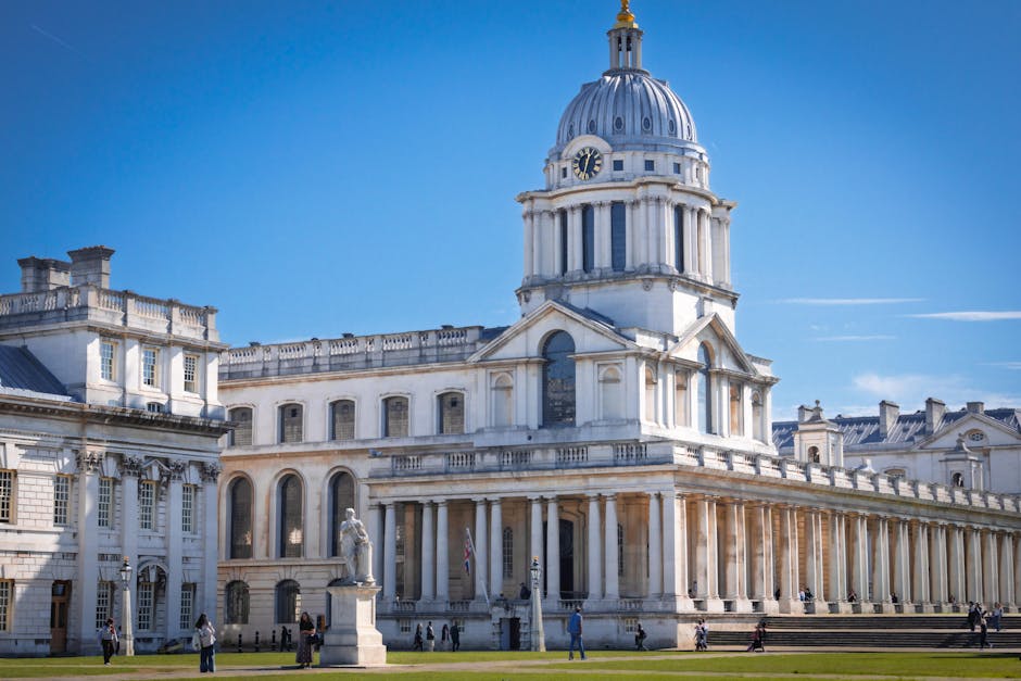 A view of a historic white stone building with classical architecture, featuring a domed clock tower at the top, surrounded by smaller statues and columns. The building is situated in an open area with a clear blue sky overhead. In front of the building, there are people walking, some near the entrance, while others are standing or sitting on the steps. The scene includes a loading area with cardboard boxes, wrapped furniture, and packing materials being carefully moved or prepared for transportation, with a van parked nearby and a person assisting with lifting and loading furniture through the doorway, indicating a home relocation or moving process. The environment is well-lit, suggesting daytime, and the overall setting reflects a professional furniture transport or house removals context as handled by [COMPANY_NAME].