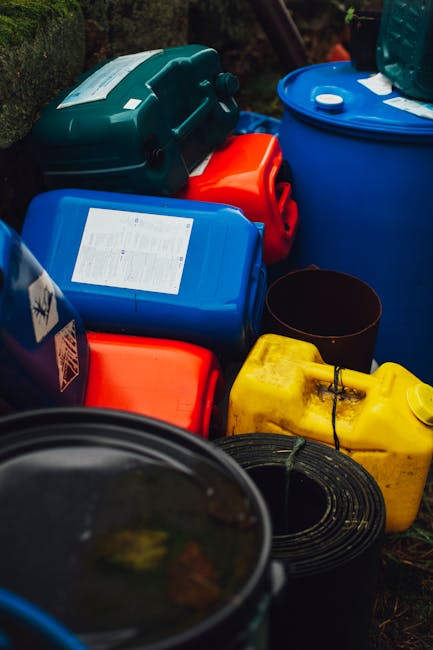 A collection of hazardous waste containers and storage drums placed outdoors on the pavement near a house, with some items stacked and others lying side by side. The containers are made of plastic in various colours including blue, yellow, and orange, with some featuring labels and caps. There is a black plastic bucket and coiled black hose among the containers. The scene appears to be part of a packing and moving process conducted by Man with Van Plumstead, with the containers arranged near the entrance of a property in preparation for transport. The image captures the outdoor environment with natural daylight, highlighting the handling and loading of potential hazardous materials during a house relocation or furniture transport.