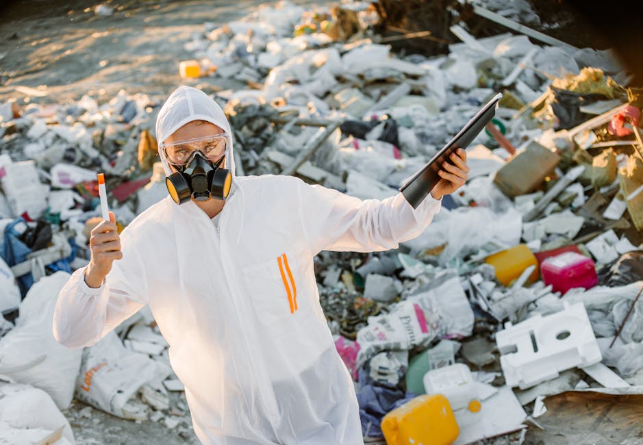 A person dressed in white protective coveralls, wearing safety goggles and a respirator mask, standing amidst a large pile of assorted household waste, including plastic containers, cardboard boxes, and packaging materials. The individual is holding a clipboard in one hand and an orange marker in the other, suggesting they are conducting an assessment or inspection related to waste removal or hazardous waste disposal as part of a home relocation or packing process. The scene takes place outdoors on a beach or shoreline, with sunlight illuminating the waste and the person. This visual highlights the importance of proper waste management during house removals, and the careful handling of hazardous materials, aligning with the services offered by Man with Van Plumstead for safe and compliant move logistics.