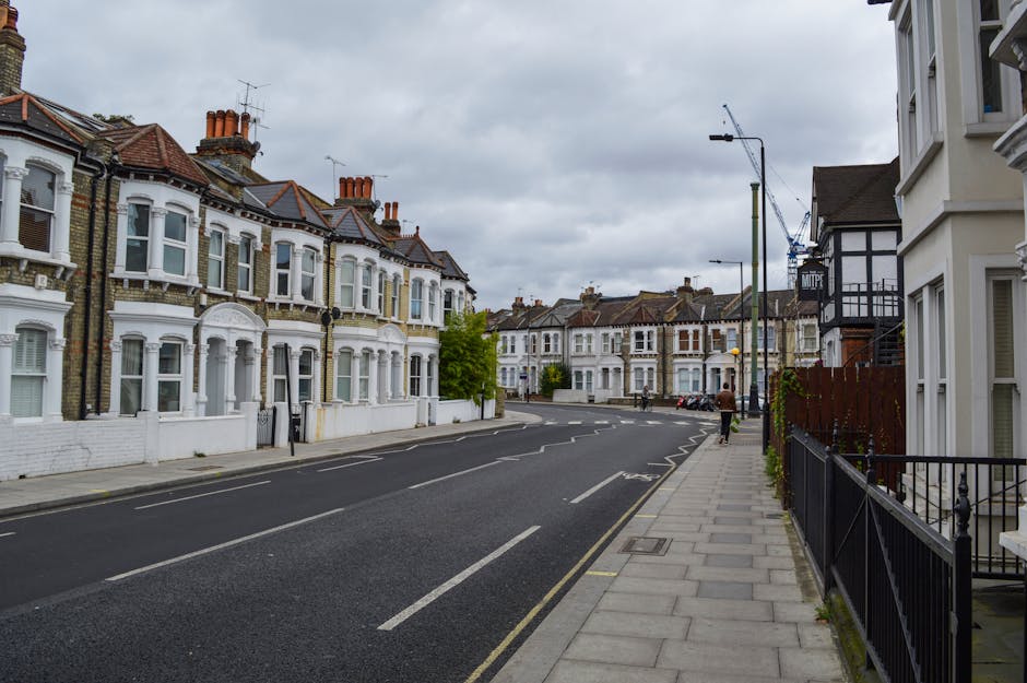 A quiet residential street lined with white and light-colored Victorian-style terraced houses, each with bay windows and brick chimneys, in Plumstead. The street features a paved sidewalk with a metal railing on the right side and a single pedestrian walking away from the camera. Parked cars are visible further down the road, which has clearly marked lanes and some parking bays. Overhead, there are streetlights and a tall crane in the background indicating ongoing construction or development. The sky is overcast with grey clouds, creating a neutral setting suitable for depicting home relocation or moving services by Man with Van Plumstead. The scene captures the typical urban environment where furniture transport, packing, and loading processes often take place during house removals in SE18, with no visible moving equipment or transport vehicles present in this particular image.