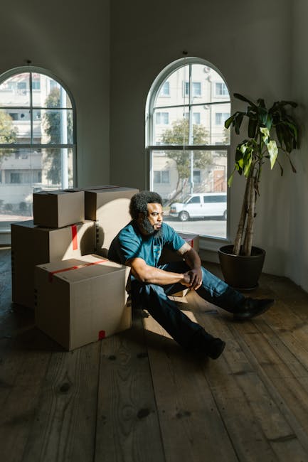 A man with curly hair and dark skin, dressed in a blue shirt and dark pants, is sitting on a wooden floor inside a room with high ceilings. Behind him, there are three large, arched windows allowing natural daylight to illuminate the space and showing a view of neighboring buildings and cars parked outside. The man is surrounded by several cardboard boxes, some closed and others partially open, with red packing straps securing them. To his right, there is a tall indoor plant in a large, black pot, adding greenery to the room. This scene captures part of a house removal process, with the man taking a moment during packing or unloading, and the environment suggests an ongoing home relocation. The setting appears tidy and well-lit, with unpacked boxes indicating active furniture transport and moving preparations, as handled by [COMPANY_NAME] and its professional removals services.