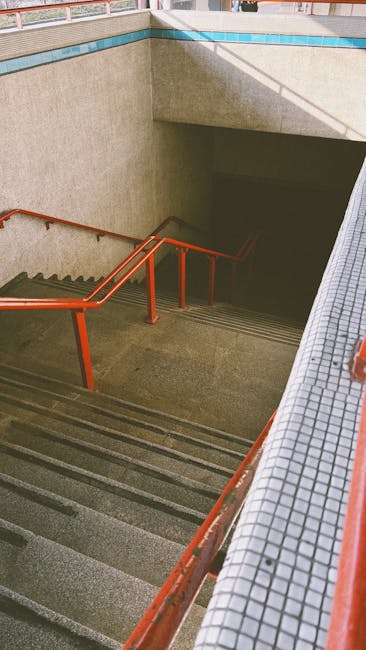 A straight staircase with yellow handrails on both sides, leading upwards inside a multi-storey industrial building or storage facility. The stairs are made of dark material with yellow edges on each step, and there are multiple steps visible, creating a clear ascending path. A white sign with black text on the middle of the staircase reads 'PLEASE KEEP LEFT.' Overhead lighting illuminates the area, and structural elements like metal beams and scaffolding are visible above, supporting the staircase and surrounding framework. The environment appears clean and organized, consistent with a professional setting involved in logistics or home relocation services. This staircase setup may be used in settings where furniture or boxes need to be transported between floors, aligning with house removal and moving operations carried out by services such as Man with Van Plumstead.