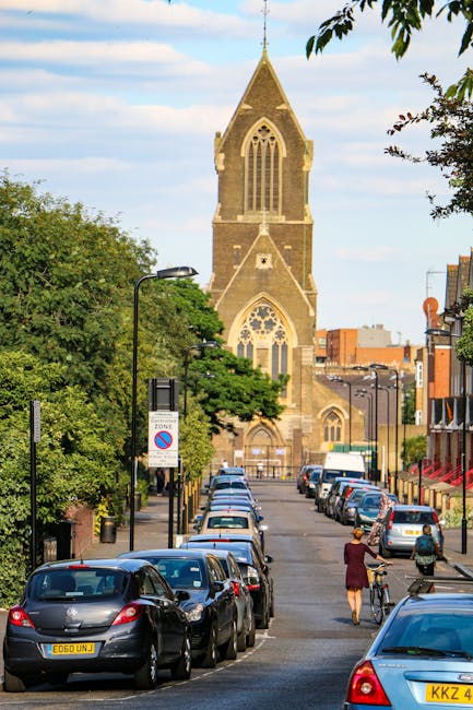 A quiet street scene on Plumstead High Street during late afternoon, with sunlight casting long shadows across the road. The street features a gentle curve with double yellow lines along the curb. A l