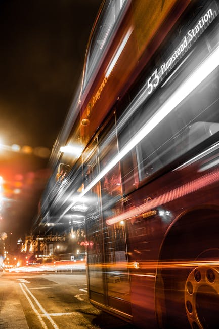 Nighttime scene outside Plumstead Station showing a red double-decker bus in motion, captured with a long exposure that creates light trails from the bus's interior and exterior lights. The bus displays route information indicating it is headed towards Plumstead Station, with blurred surrounding streetlights and vehicle lights emphasizing the busy urban environment. The pavement and roadside are faintly visible, with reflections of streetlights on wet surfaces. The image highlights public transportation in the vicinity of Plumstead, relevant to residents or professionals involved in home relocation or moving logistics, such as those coordinated by Man with Van Plumstead, especially during busy periods.