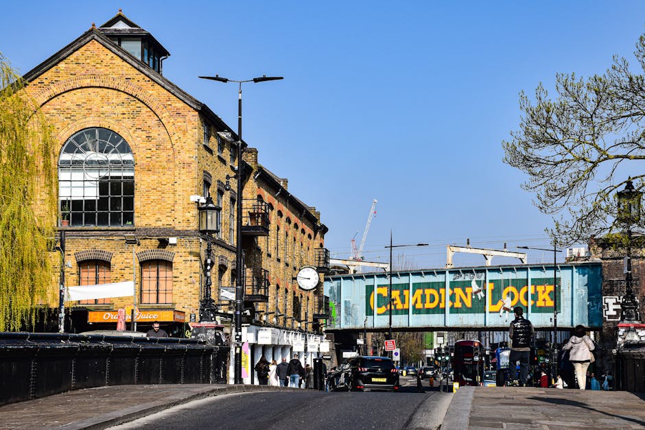 A street scene featuring a large brick building with arched windows and a rounded roof, situated next to a railway bridge with a blue cargo container labeled 'Caidin Lock' crossing above. The scene is set during daytime with a clear blue sky, and several pedestrians walking on the pavement near parked cars and delivery trucks. Visible elements include a street clock, lampposts, and sporadic trees with budding leaves. The environment suggests an urban area with active transportation and loading activities, indicating a busy environment suitable for house removals and furniture transport. In the context of home relocation or packing and moving services, this image highlights the typical setting where Man with Van Plumstead might operate during client house moves or logistical operations in a vibrant city location.